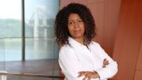 A woman with curly hair in a white shirt, arms crossed, standing indoors with a bridge visible in the background